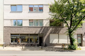 Un immeuble à deux étages abritant un café au rez-de-chaussée, avec des places assises en plein air et un arbre devant.