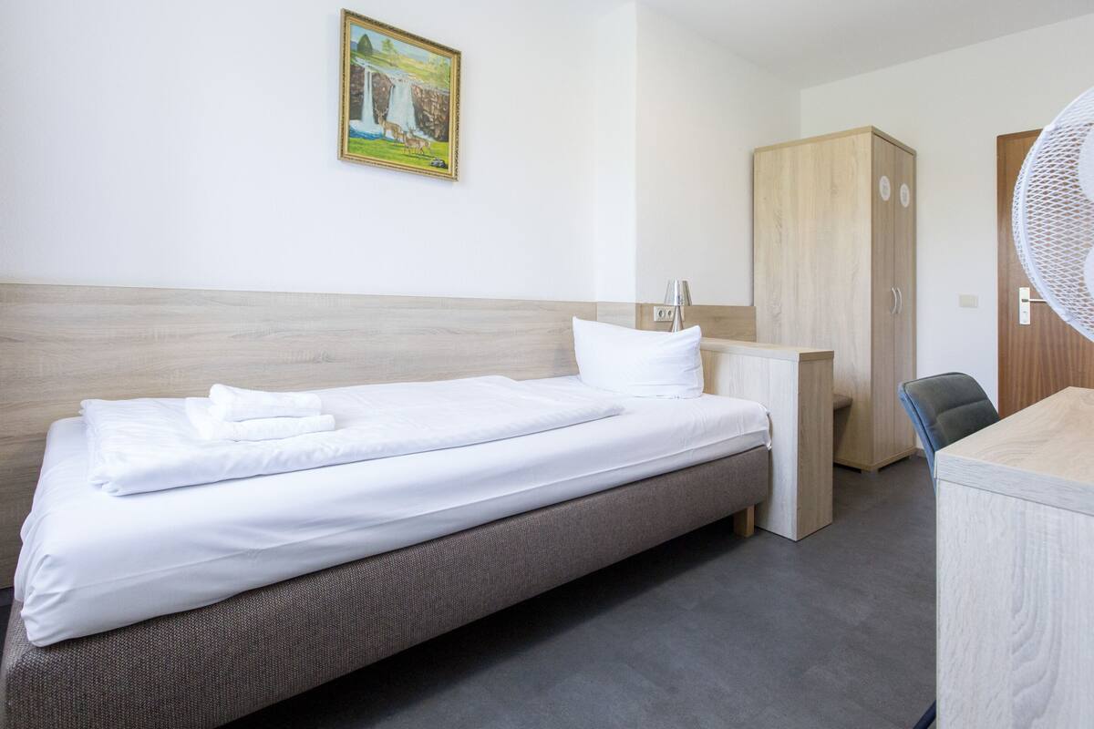 A neatly made bed with white linens, a wooden headboard, a small desk with a chair, a wardrobe, and a wall-mounted fan.