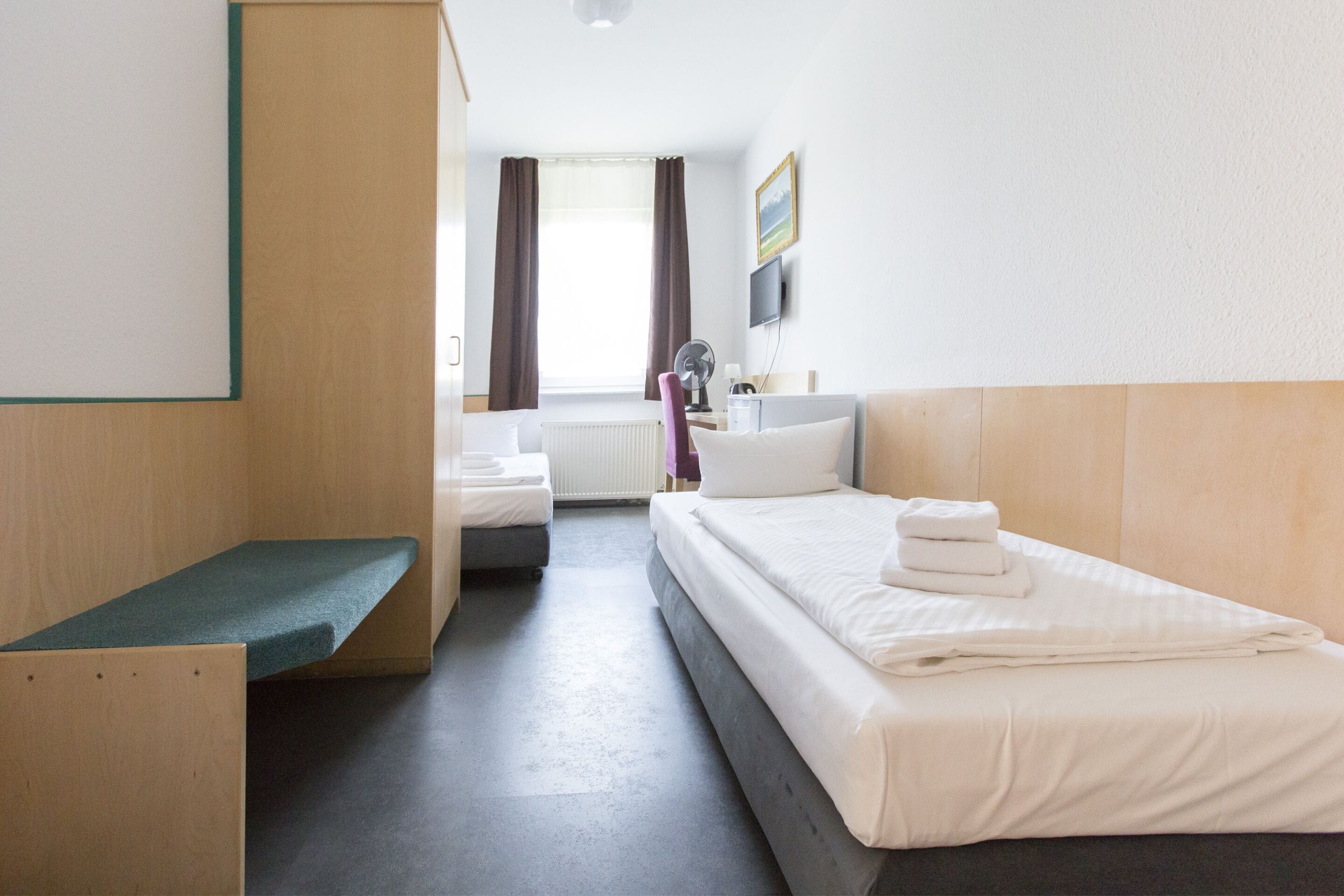 A hotel room with a bed, a bench, a desk, a chair, a TV, and a window with curtains.