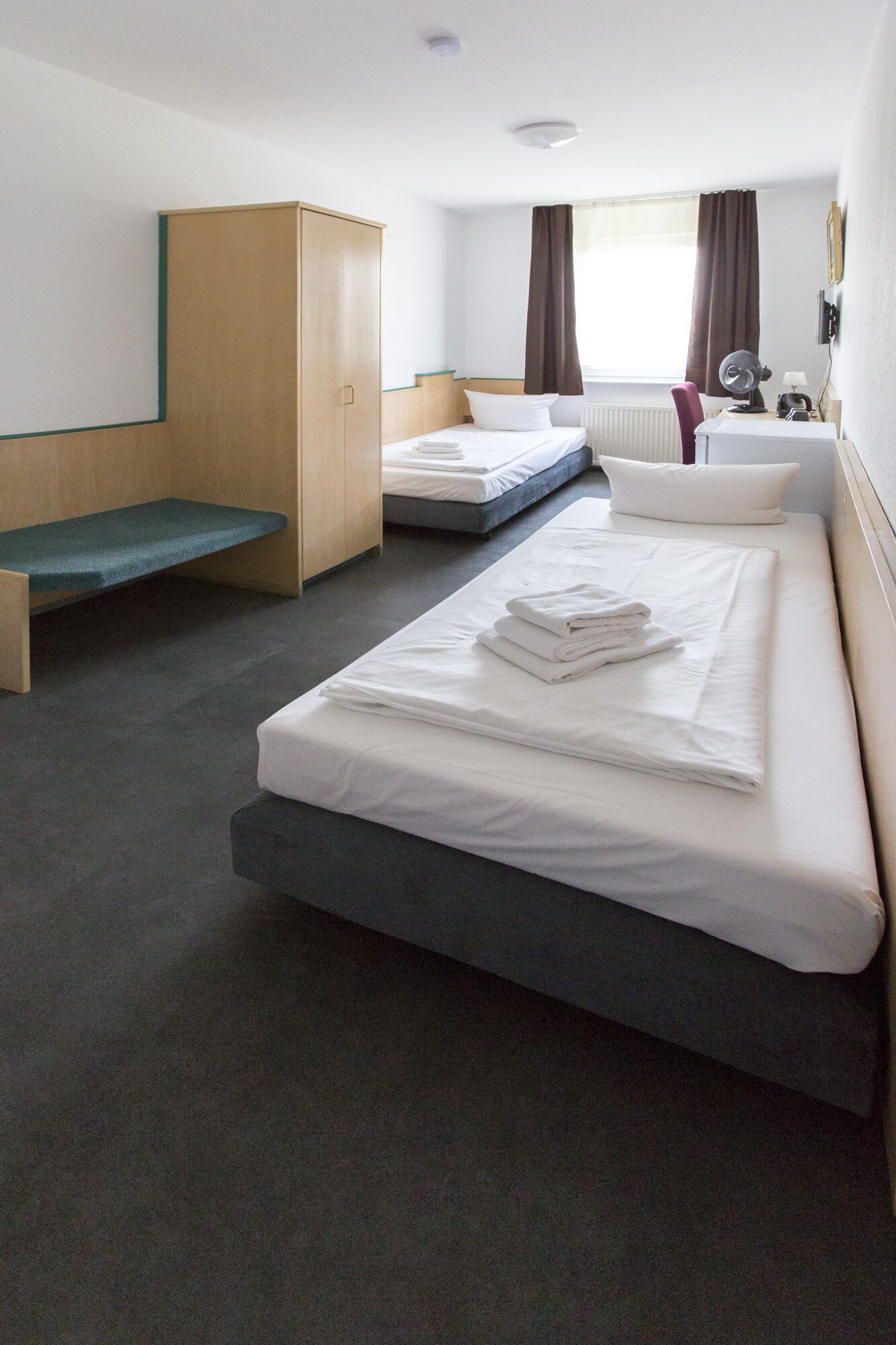 Ein Hotelzimmer mit einem Einzelbett, einem Schreibtisch, einem Stuhl, einem Kleiderschrank und einem Fenster mit Vorhängen.