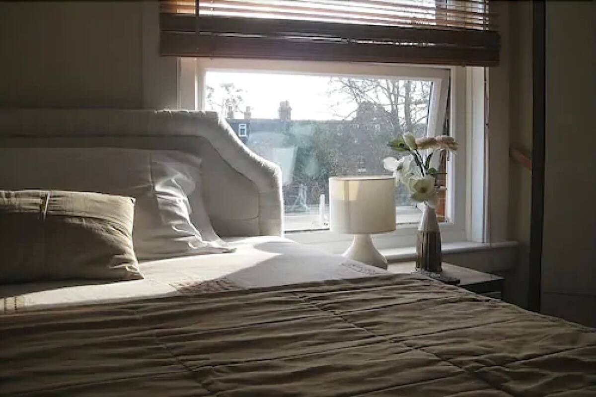 A bedroom with a bed, pillows, a bedside table with a lamp, and a window with a view of buildings and trees.