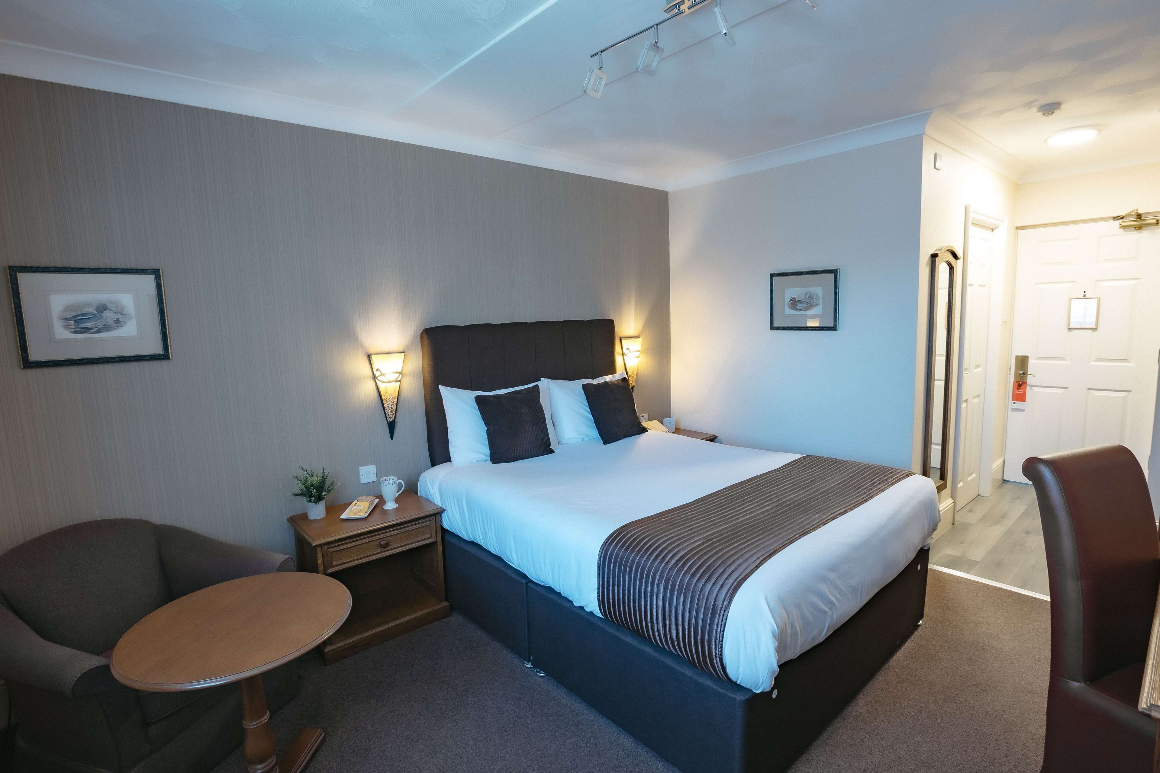 A hotel room with a large bed, bedside tables, a small round table, two chairs, and framed pictures on the walls.