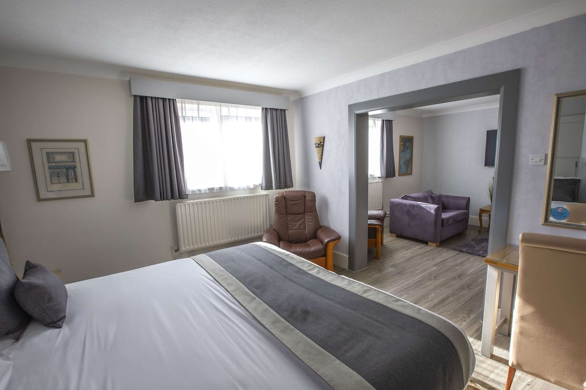 A hotel room with a large bed, a brown leather chair, a purple armchair, and a mirror on the wall.