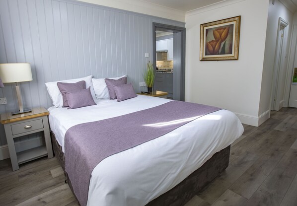 A neatly arranged bedroom with a large bed, a bedside table with a lamp, and a view of the kitchen through an open door.