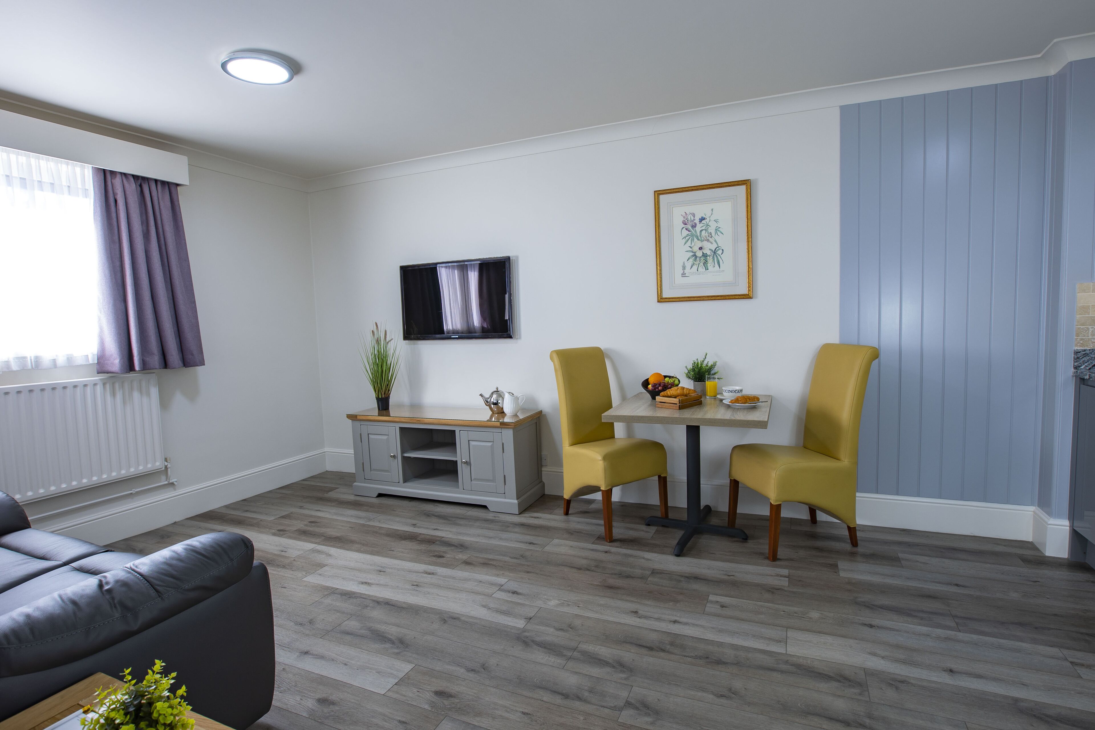 Un salon moderne avec un canapé gris, une table à manger accompagnée de deux chaises jaunes, un meuble gris, un téléviseur à écran plat et une estampe botanique encadrée accrochée au mur.