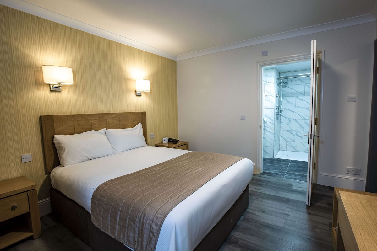 A hotel room with a wooden headboard, a large bed with white linens, two bedside lamps, a wooden nightstand, and a bathroom visible through an open door.