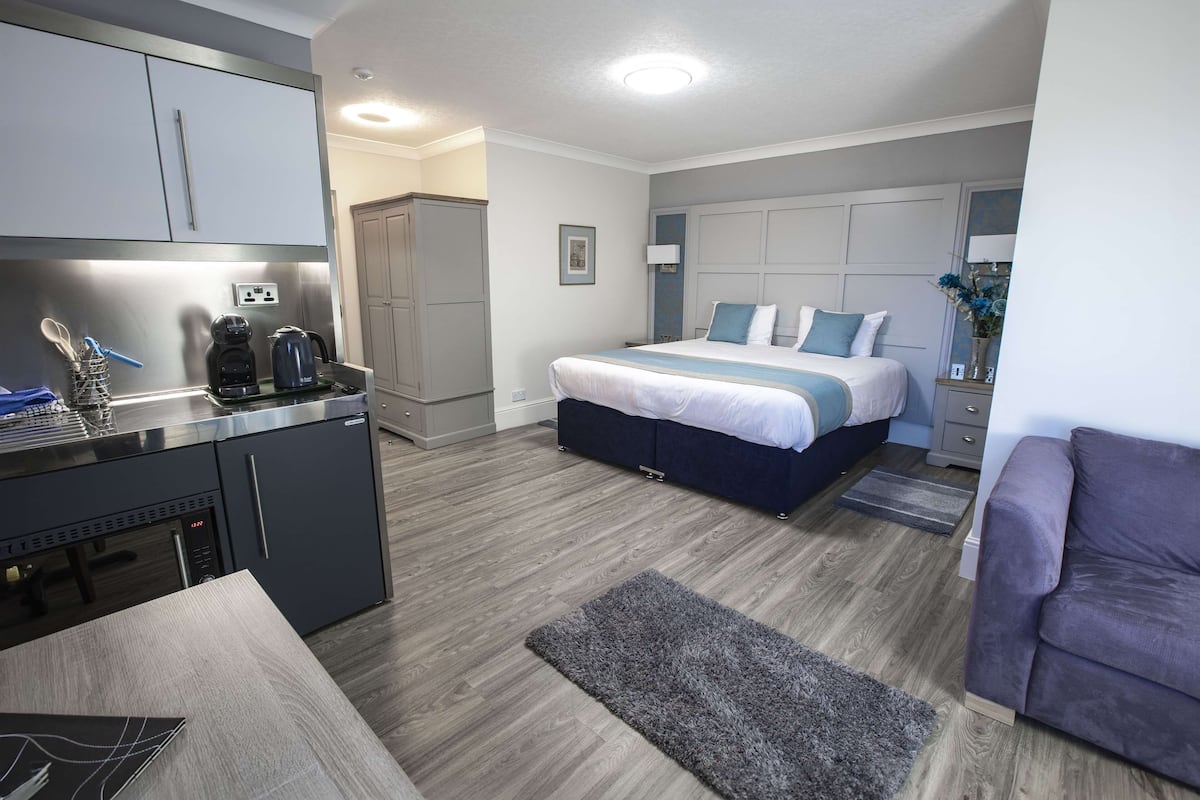 A modern hotel room with a kitchenette, a bed with blue and white bedding, a grey sofa, and a grey rug.