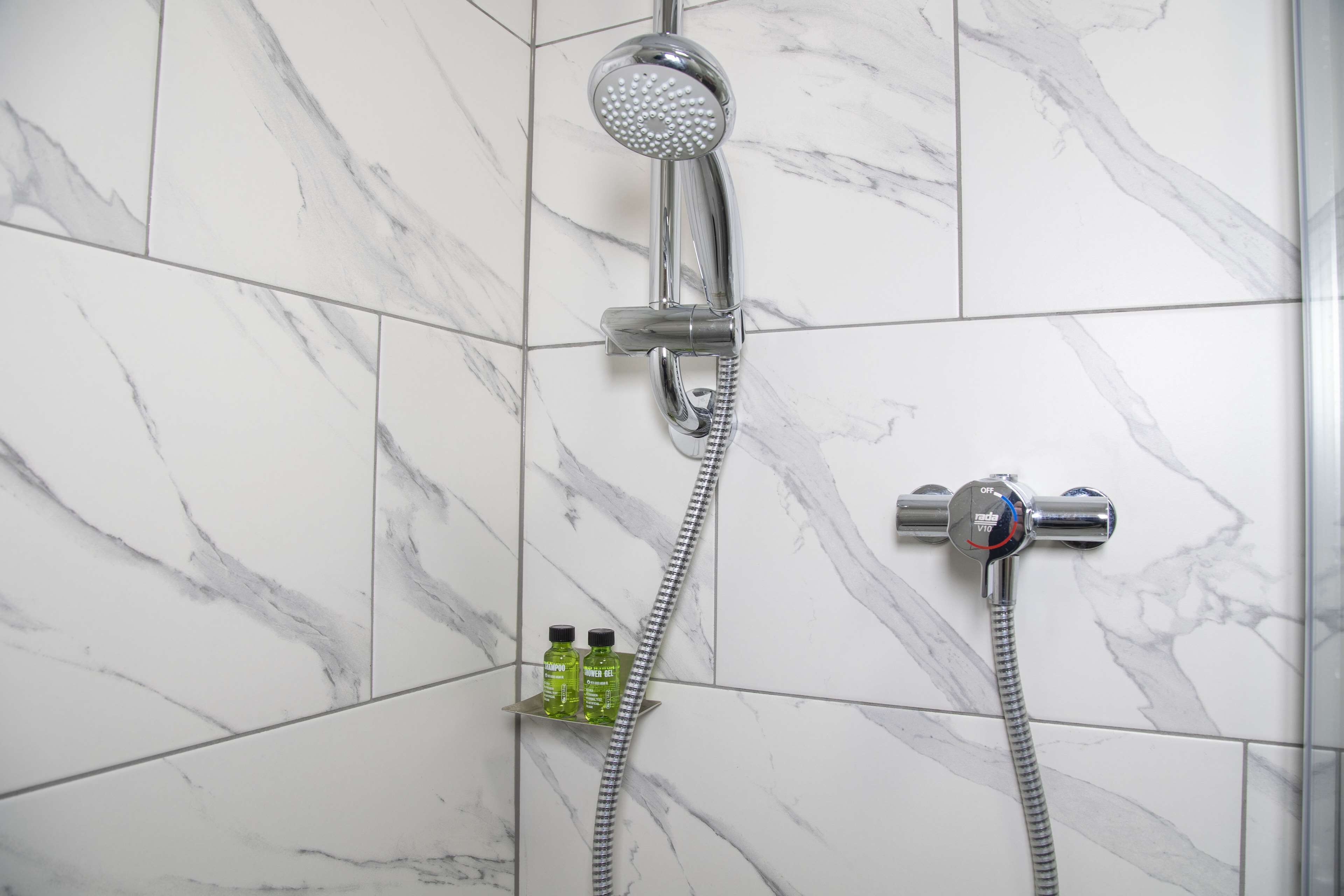 A bathroom with a shower, a handheld showerhead, and a handheld faucet. There are two bottles on a shelf below the shower.