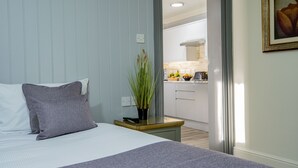 A bedroom with a bed, pillows, a bedside table with a plant, and a view into a kitchen area. - Best Western Manor Hotel (Gravesend)