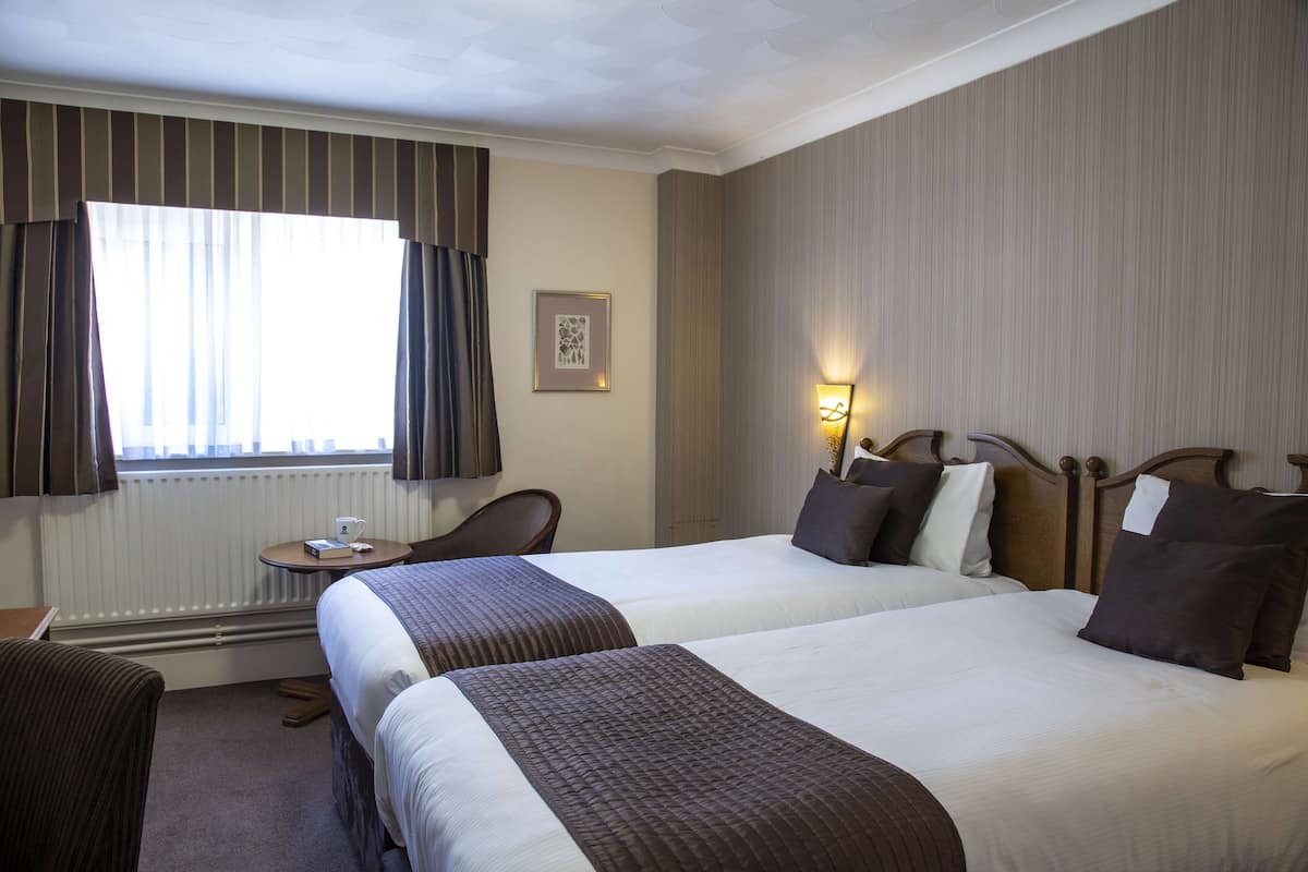 A hotel room with two beds, a small table with a cup, a chair, and a radiator.