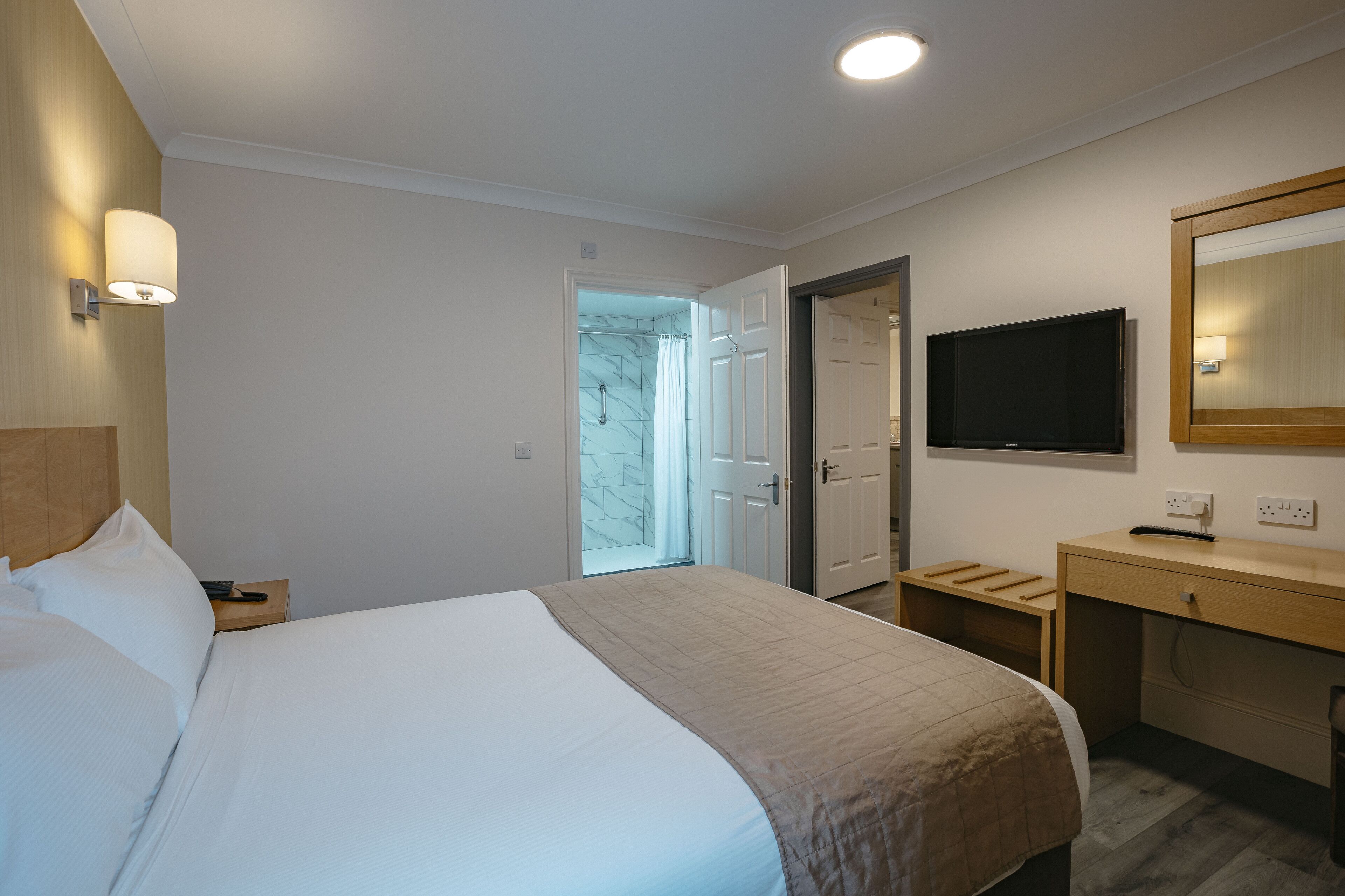 Quarto de hotel com cama, mesa de cabeceira, escrivaninha, televisão e porta do banheiro.
