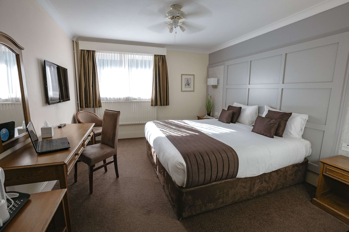 A hotel room with a large bed, a desk with a laptop, a chair, a bedside table, and a ceiling fan.