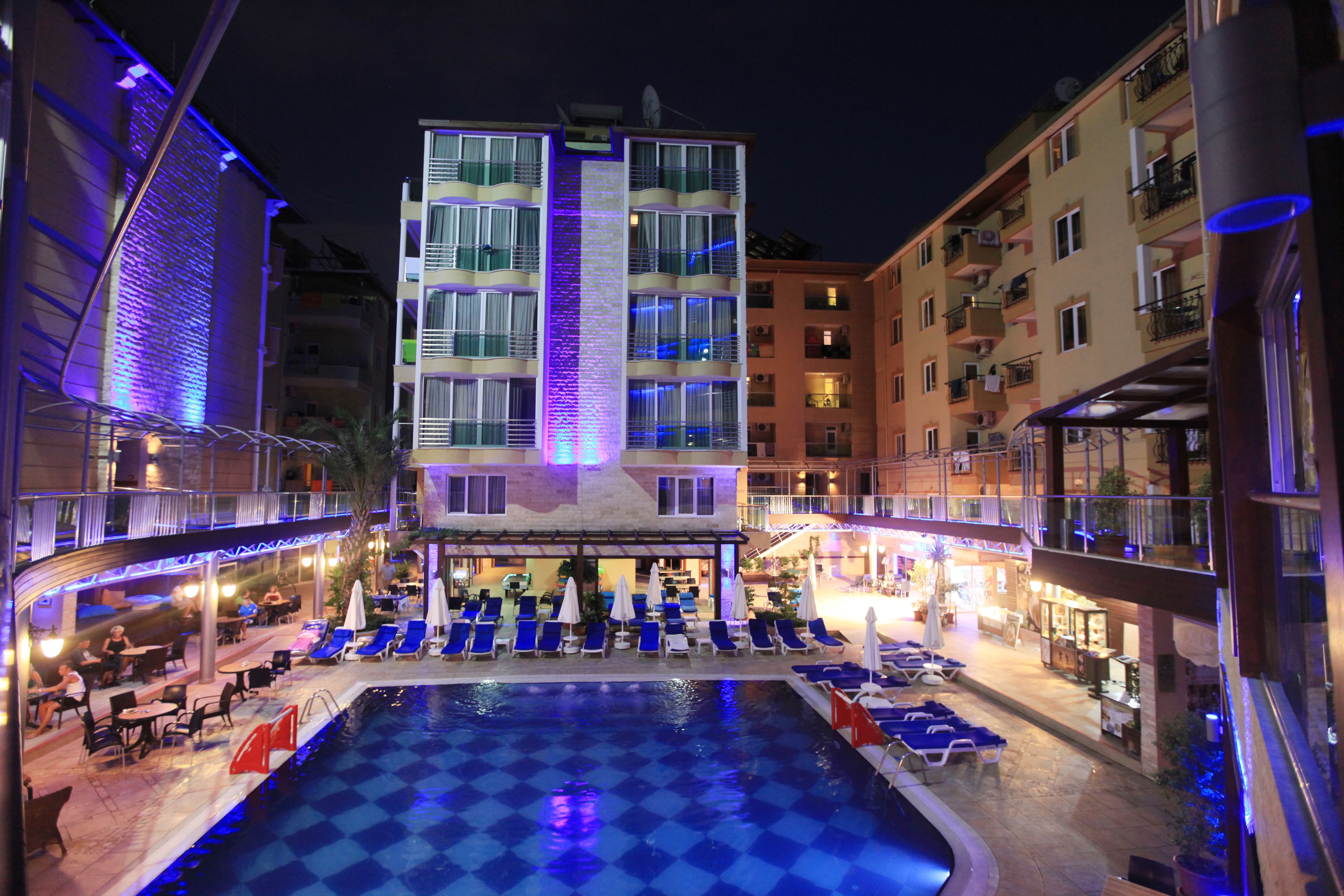 Un espace piscine d’hôtel avec des chaises longues et des parasols, entouré d’immeubles à plusieurs étages dotés de balcons.