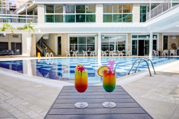 Zona de piscina con dos cócteles coloridos sobre una mesa, una piscina y un edificio con asientos al aire libre y una escalera.