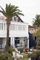 Un edificio blanco con balcón, palmeras y un jardín con plantas en macetas y flores.