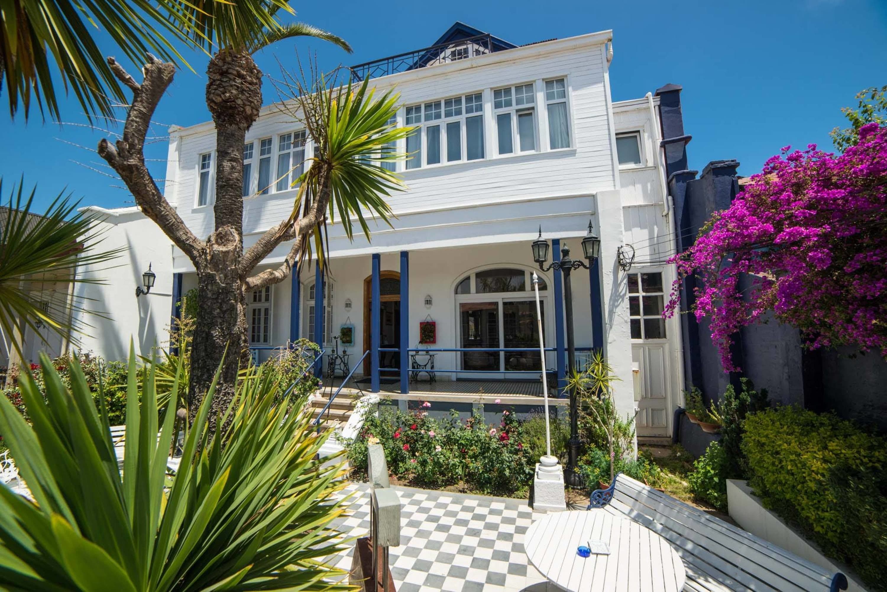 A two-story white building with a balcony, surrounded by lush greenery and a checkered outdoor area.