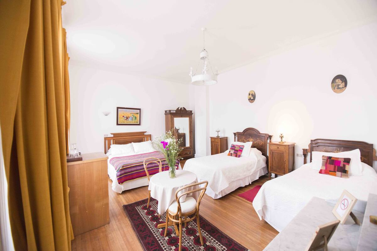 A hotel room with two beds, a wooden headboard, a bedside table with a lamp, a small table with a chair, and a red rug.