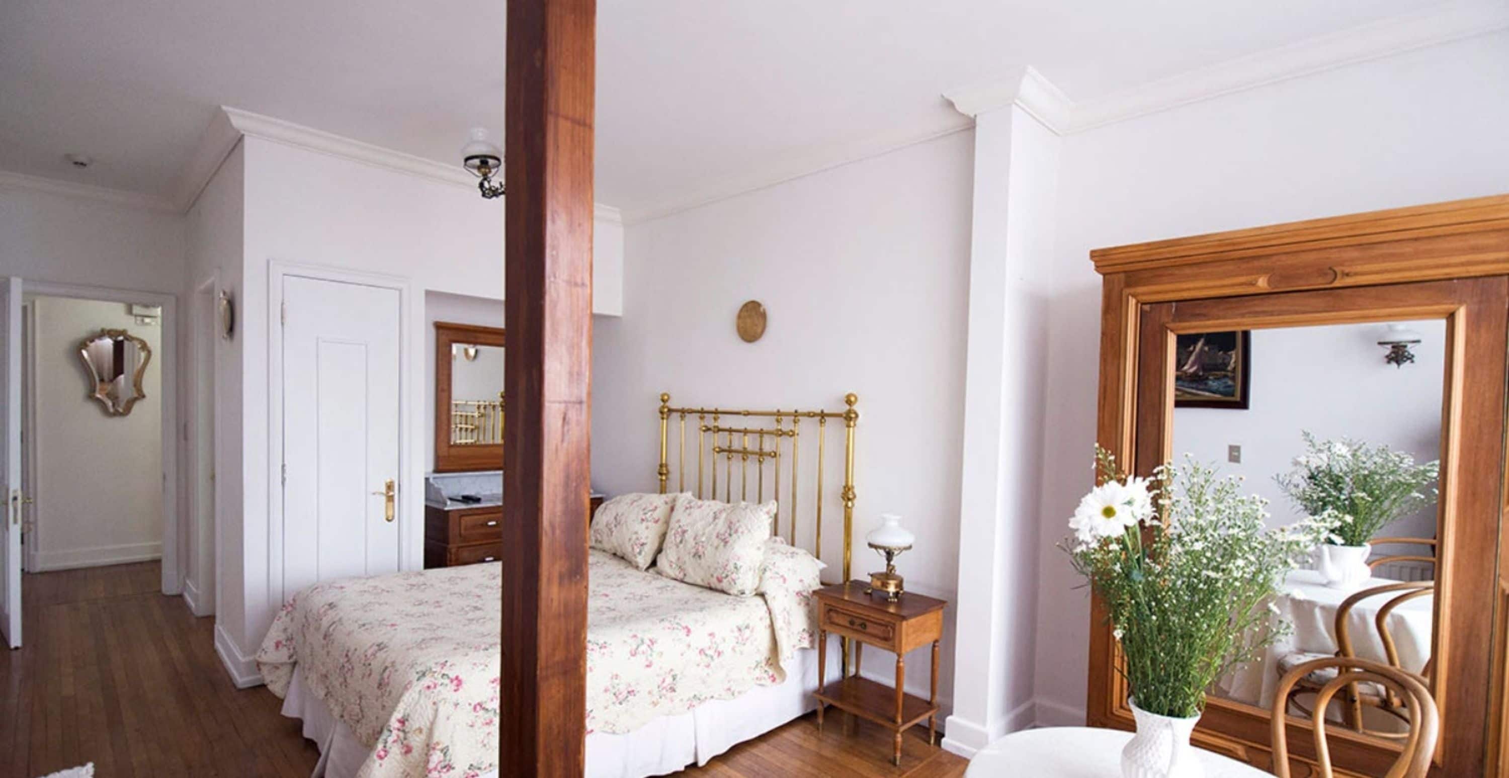 Quarto com estrutura de cama de madeira, mesa de cabeceira, espelho e arranjo floral.