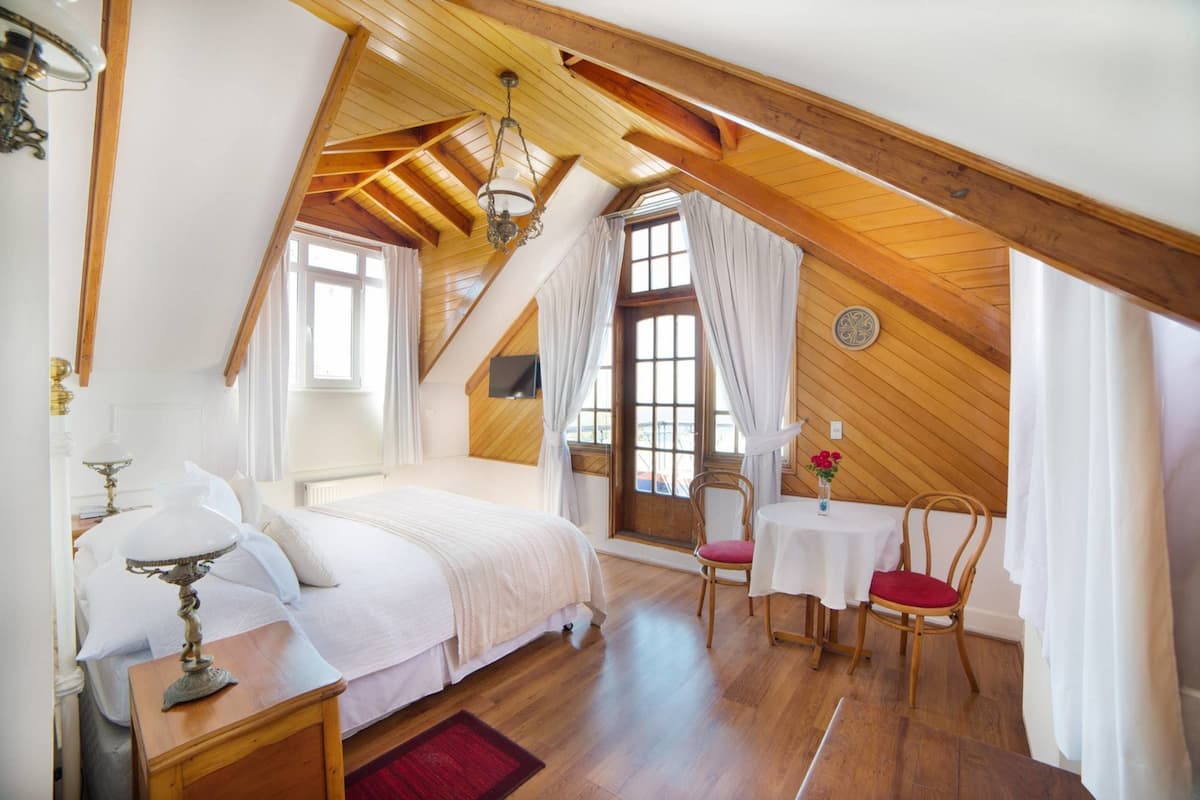 A bedroom with a sloped ceiling, wooden beams, a bed, a table with chairs, and a window with curtains.
