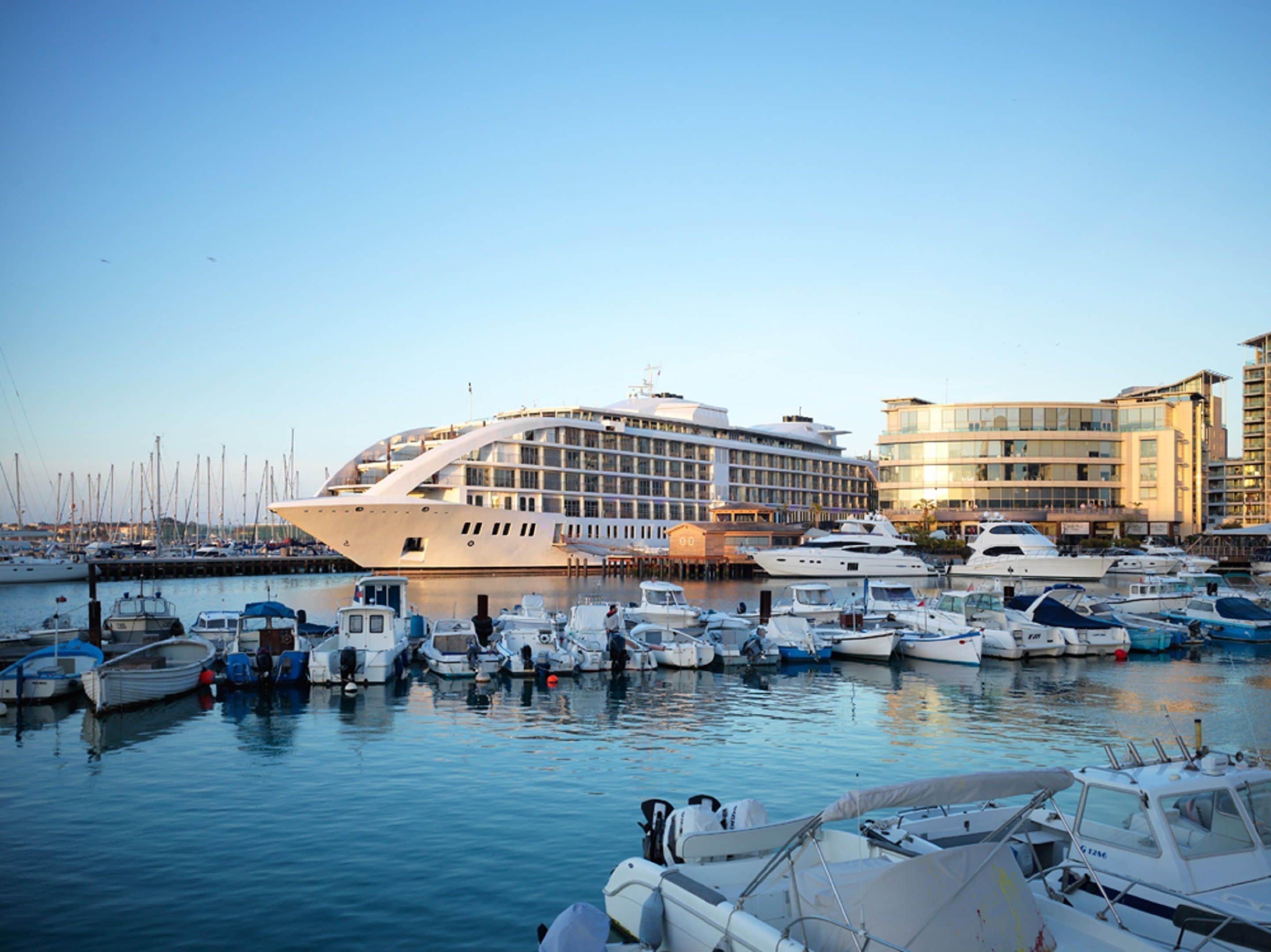 A marina with various boats docked, a large cruise ship, and modern buildings in the background.