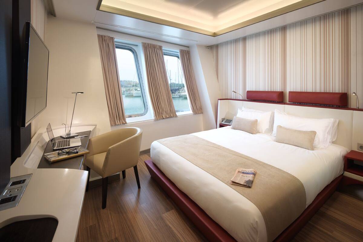 Ein modernes Hotelzimmer mit einem großen Bett, einem Schreibtisch mit Laptop, einem Sessel und einem Fenster mit Vorhängen.