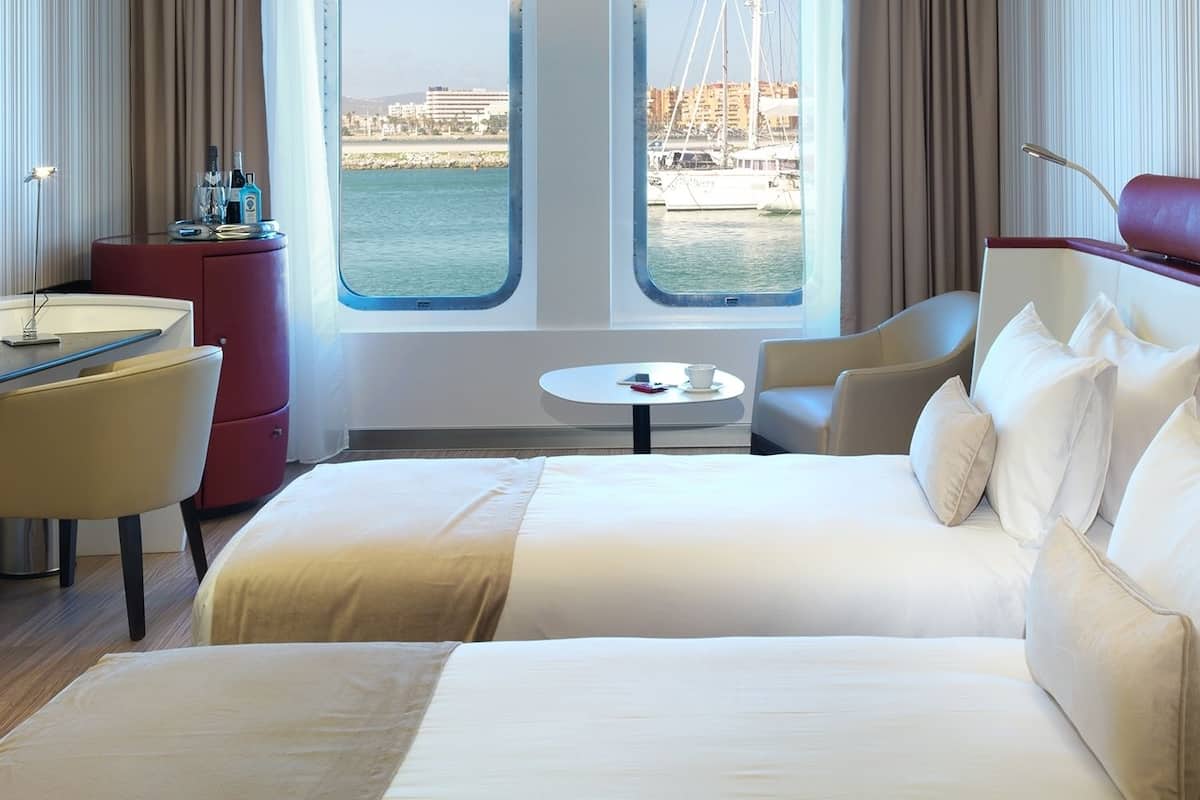Ein Hotelzimmer mit Bett, Schreibtisch, Stuhl, einem kleinen Tisch und Blick auf das Meer und die Gebäude.