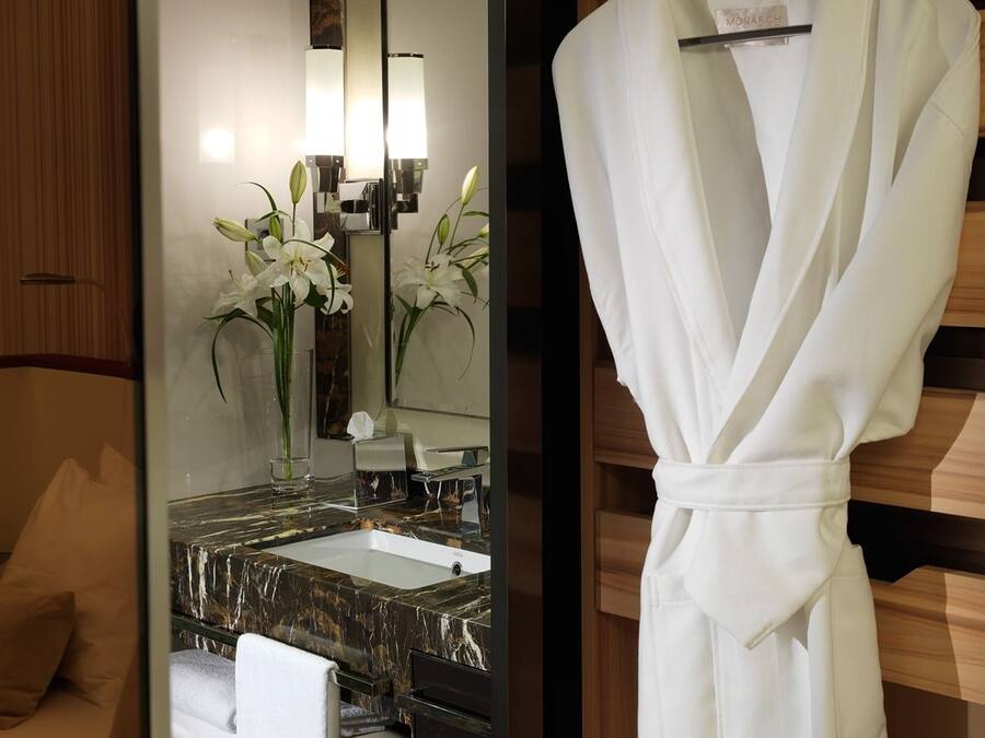 Un baño de hotel con un lavamanos de mármol, una bata blanca colgada en un perchero y un espejo con una lámpara empotrada en la pared.