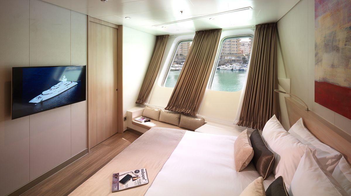 A modern hotel room with a large bed, a TV, a sofa, and a view of the water through windows.