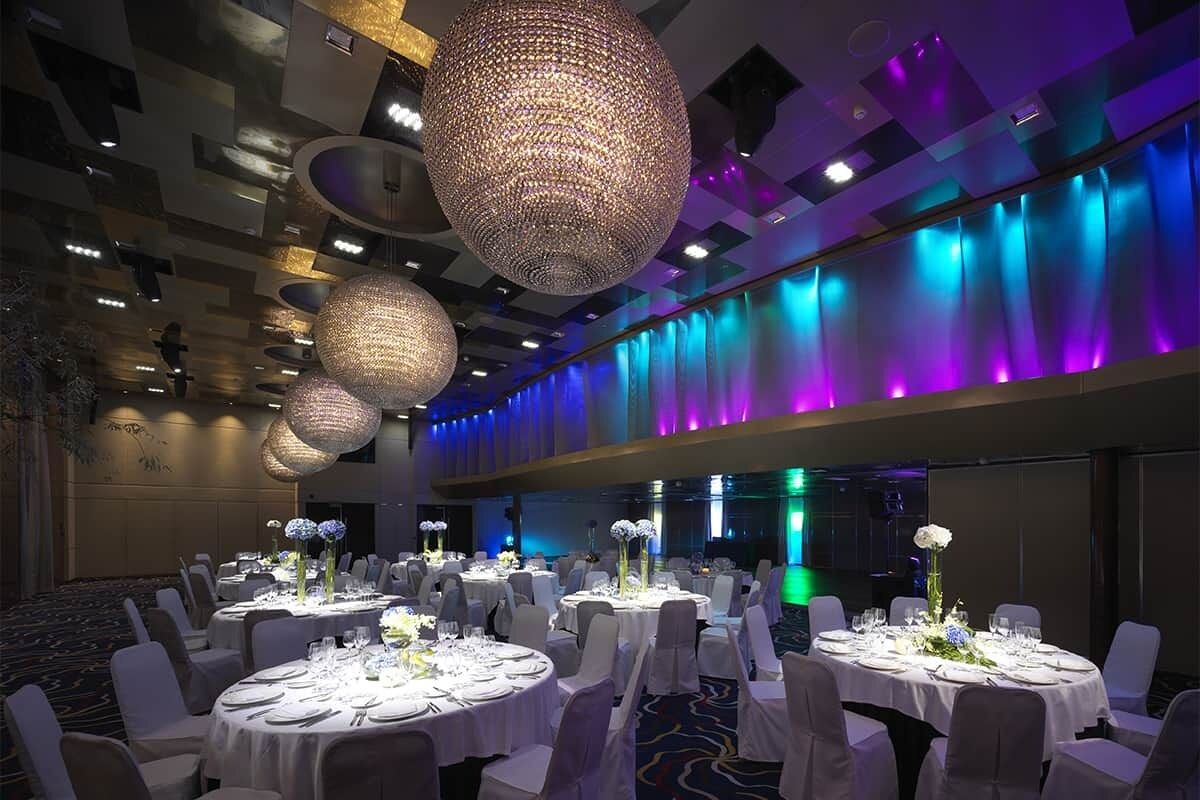 A banquet hall with round tables set for dining, large hanging chandeliers, and a modern ceiling design.