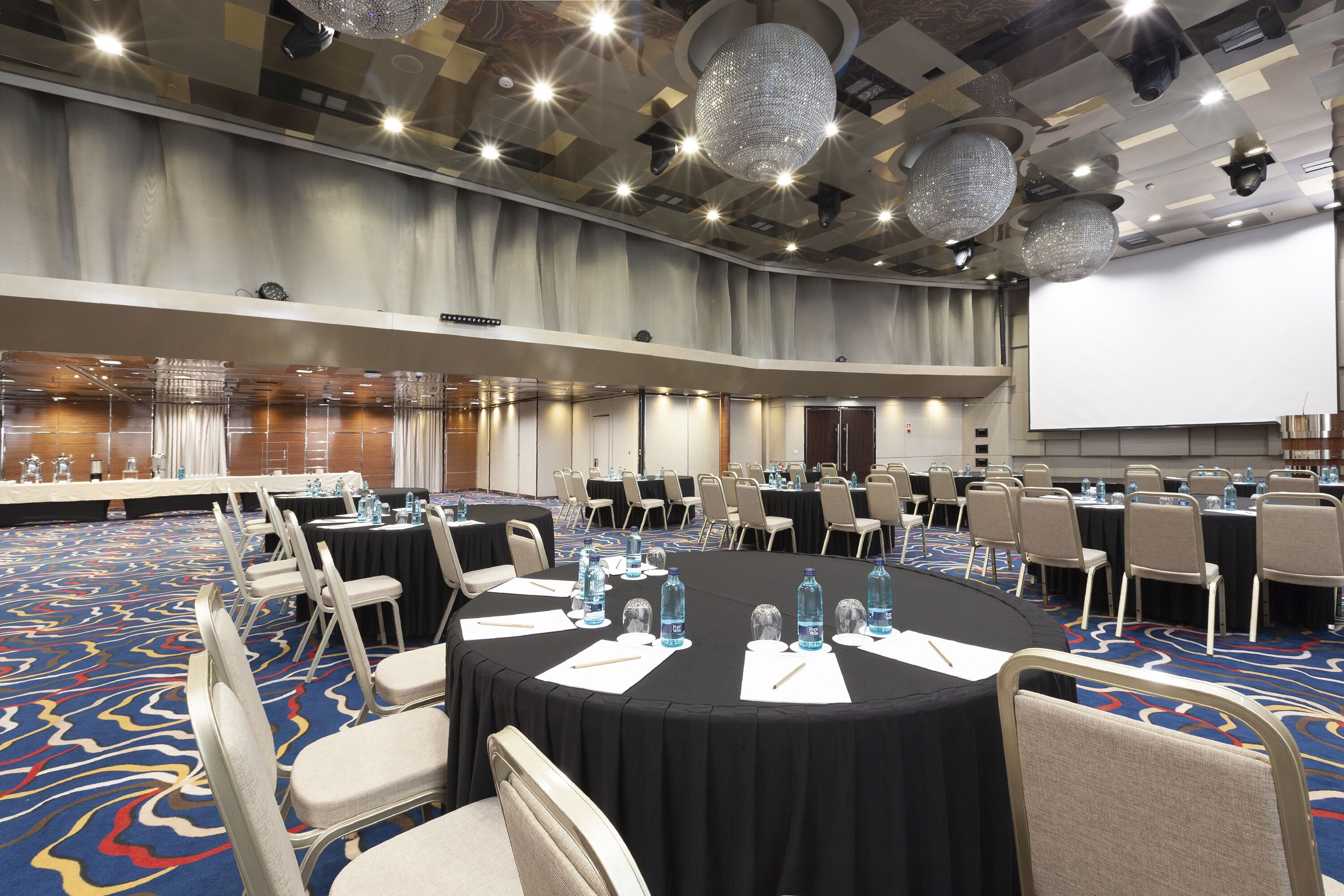 Un salón de conferencias con mesas redondas, sillas, botellas de agua y una pantalla de proyector.