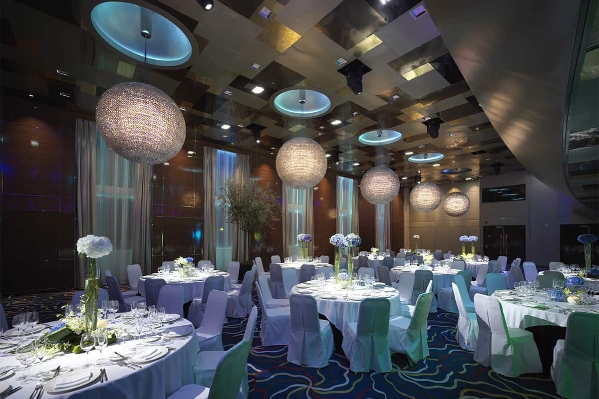 Un salón de banquetes con mesas redondas dispuestas para cenar, grandes lámparas esféricas colgantes y un diseño de techo moderno.