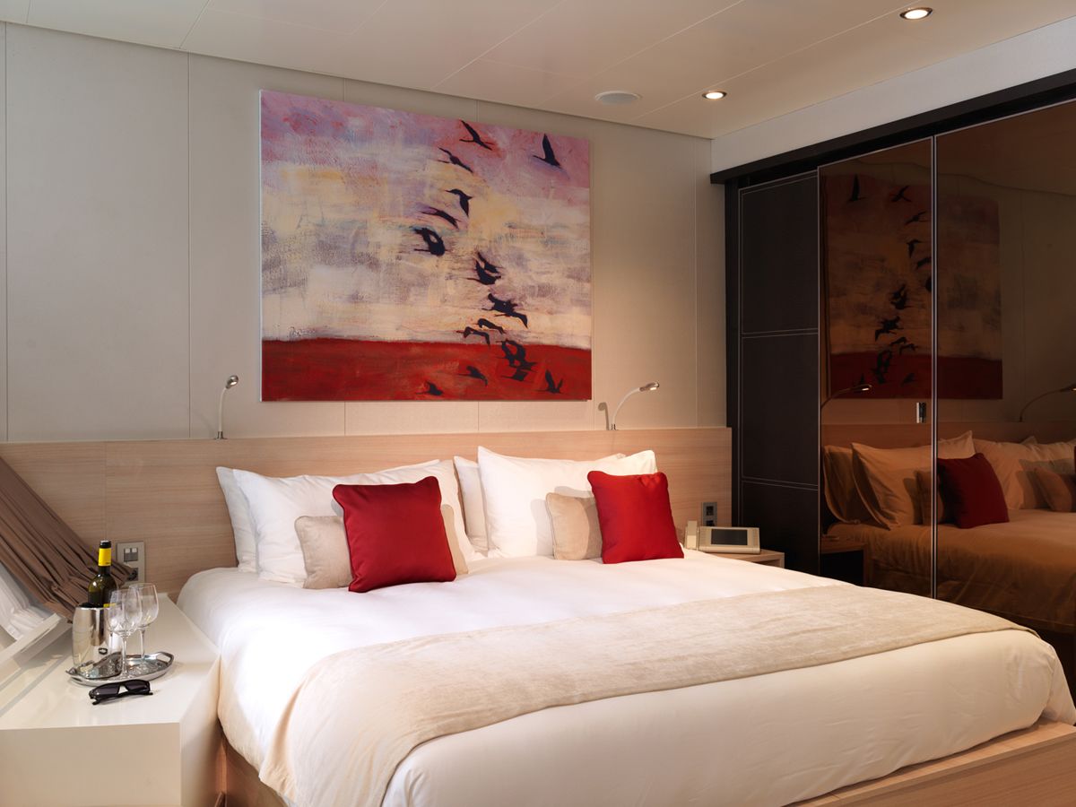 A modern hotel room with a large bed, red pillows, a bedside table with a wine bottle and glasses, and a large abstract painting above the bed.