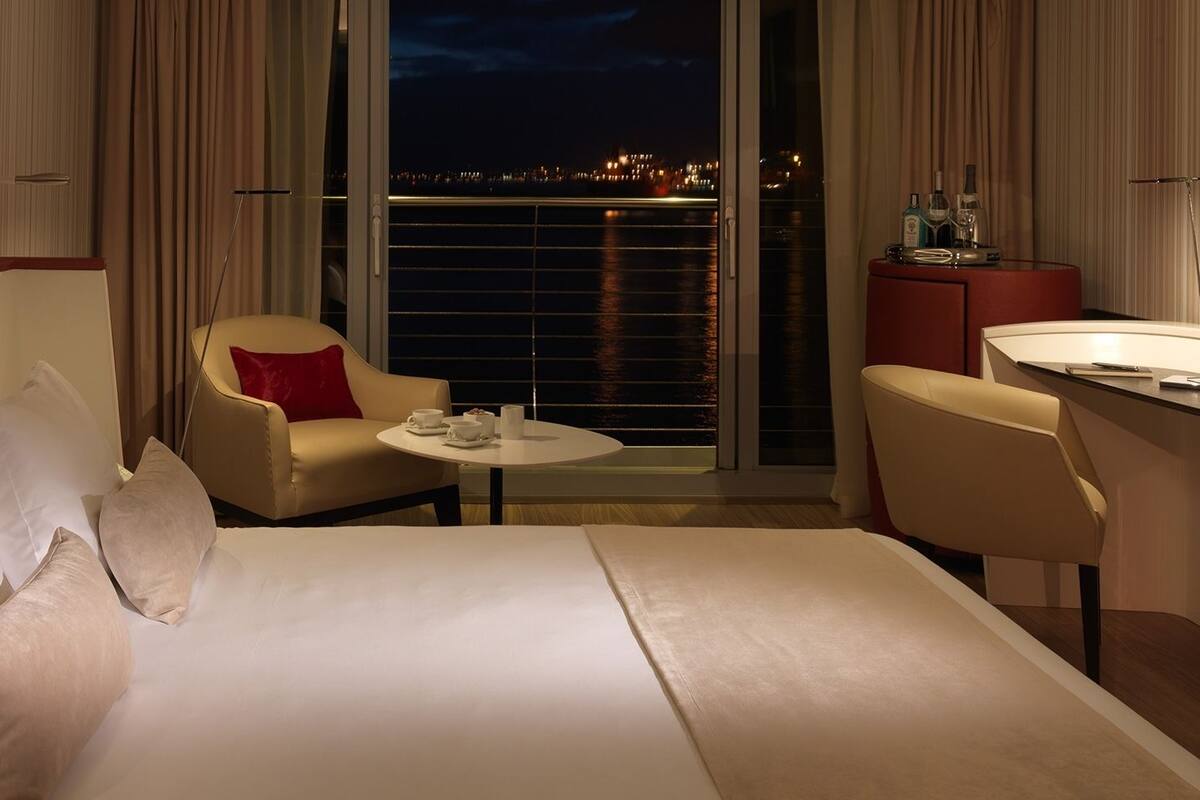 Ein Hotelzimmer mit Bett, Stuhl, kleinem Tisch und Schreibtisch mit Blick auf die beleuchtete Stadt.