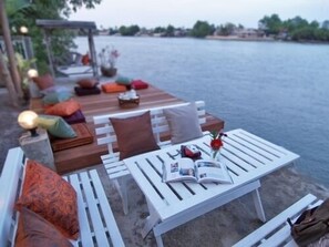 A waterfront outdoor seating area with white tables and benches, cushions, and a book. - The Legend Maeklong (Samut Songkhram)