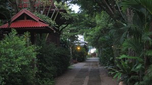A pathway leading to a traditional red-roofed building surrounded by lush greenery. - The Legend Maeklong (Samut Songkhram)