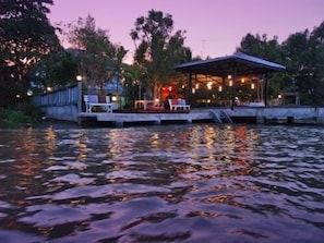 A riverside restaurant with outdoor seating and a covered area, surrounded by trees and a calm water body. - The Legend Maeklong (Samut Songkhram)