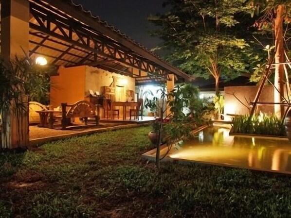 Un espacio exterior bien iluminado con una pérgola de madera, un pequeño estanque y una zona de estar con mesas y sillas.