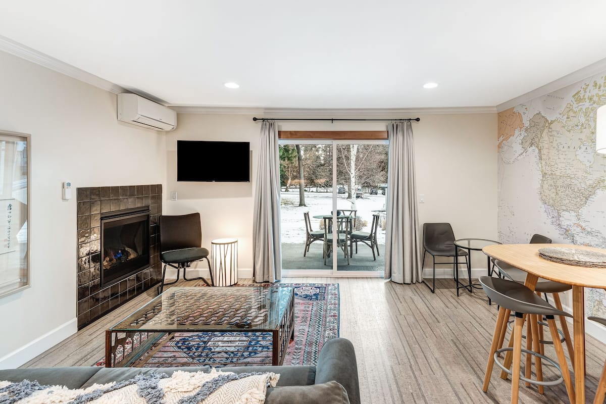 A modern living room with a fireplace, a flat-screen TV, a dining area with a map on the wall, and a view of the outside.