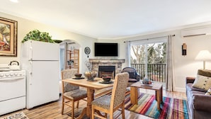 A well-lit living room with a fireplace, a dining area with a table and chairs, a kitchen with a refrigerator and stove, and a balcony with a view of trees. - Pioneer Park Rentals (Bend)
