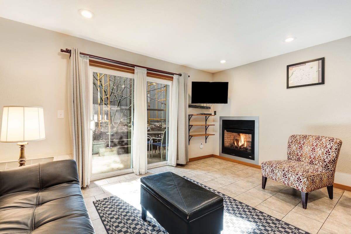 A living room with a sofa, a patterned armchair, a black ottoman, a fireplace, and a TV mounted on the wall.