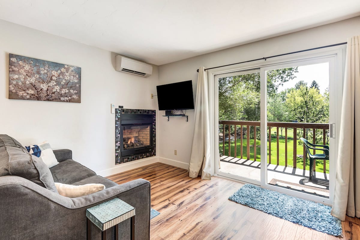 A living room with a fireplace, a sofa, a television, and a balcony with a view of the outdoors.