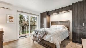 A bedroom with a large bed, wooden flooring, a sliding glass door to a balcony, and dark wood cabinetry. - Pioneer Park Rentals (Bend)