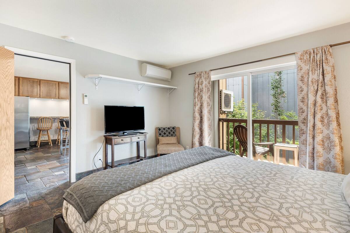 A hotel room with a bed, a TV, a desk, and a balcony with a view of greenery.