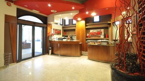 A reception area with a curved wooden desk, a large potted plant, and a glass door leading outside. - Planet Hotel (Maranello)