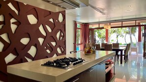 Una cocina moderna con una isla central, una zona de comedor con sillas de mimbre y ventanas grandes con cortinas.