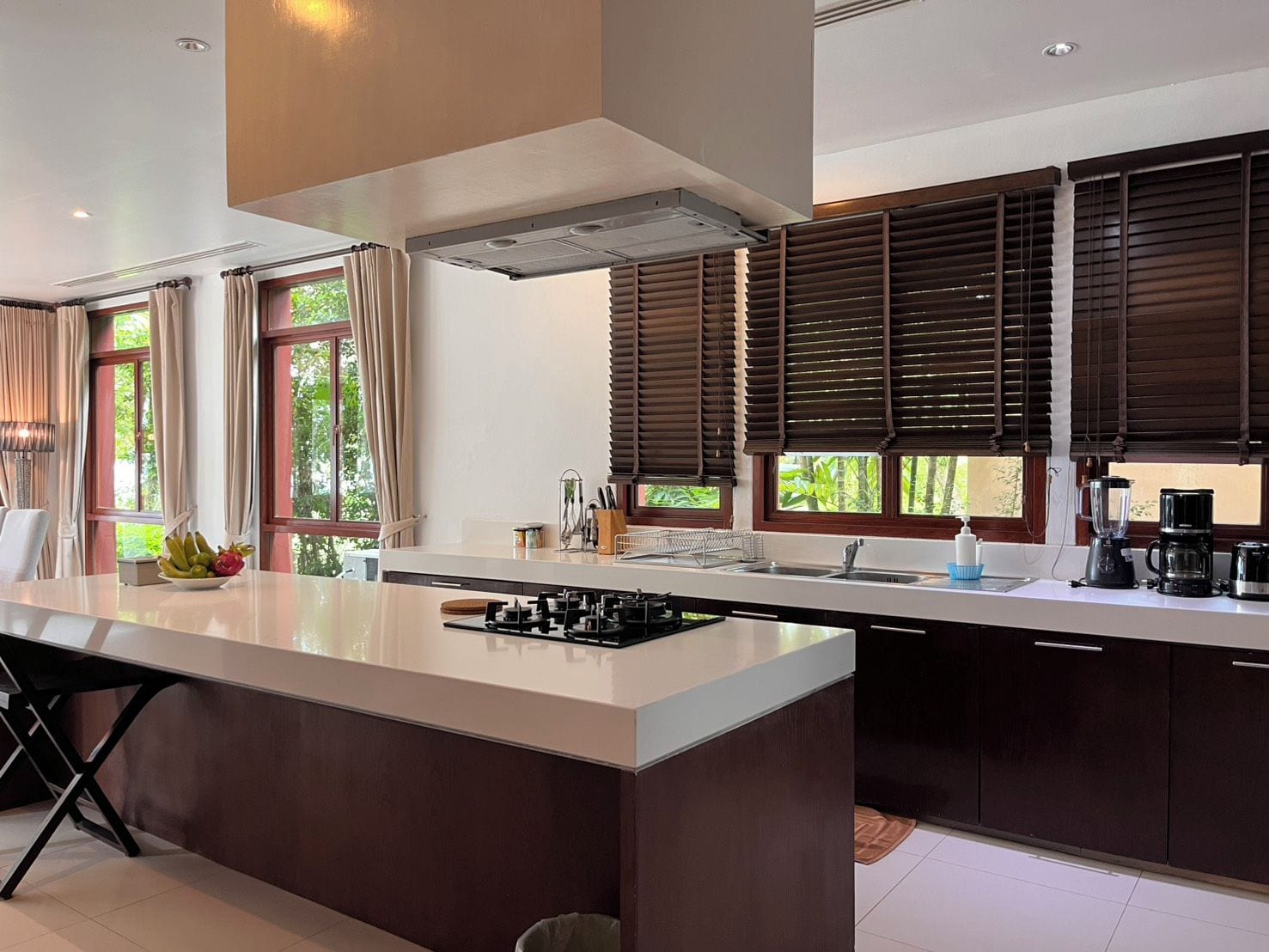 Una cocina moderna con una isla central, muebles oscuros y un amplio ventanal con persianas.