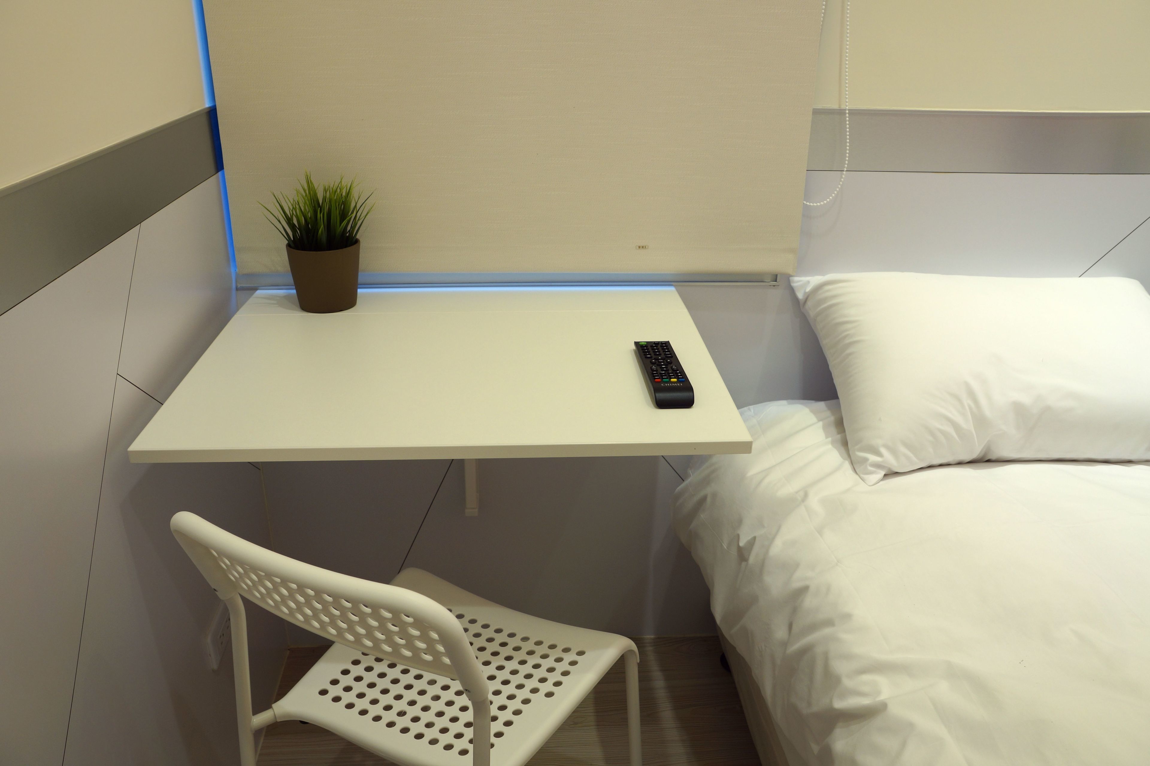 A small, neatly arranged bedroom with a single bed, a bedside table with a remote control, a potted plant, and a white chair.