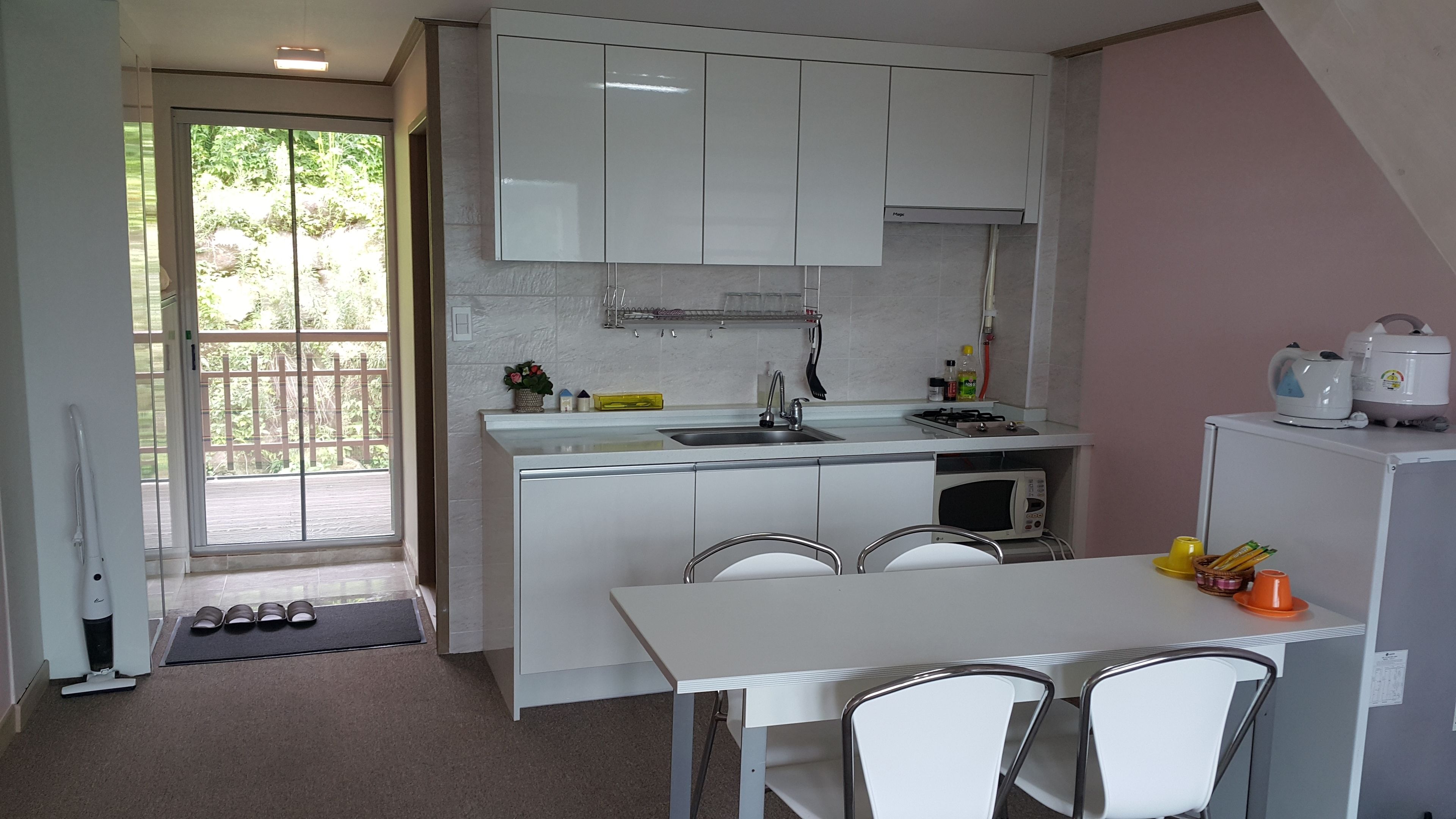 A compact kitchen with white cabinets, a sink, a microwave, and a small refrigerator. There is a dining table with chairs and a window providing a view of the outdoors.
