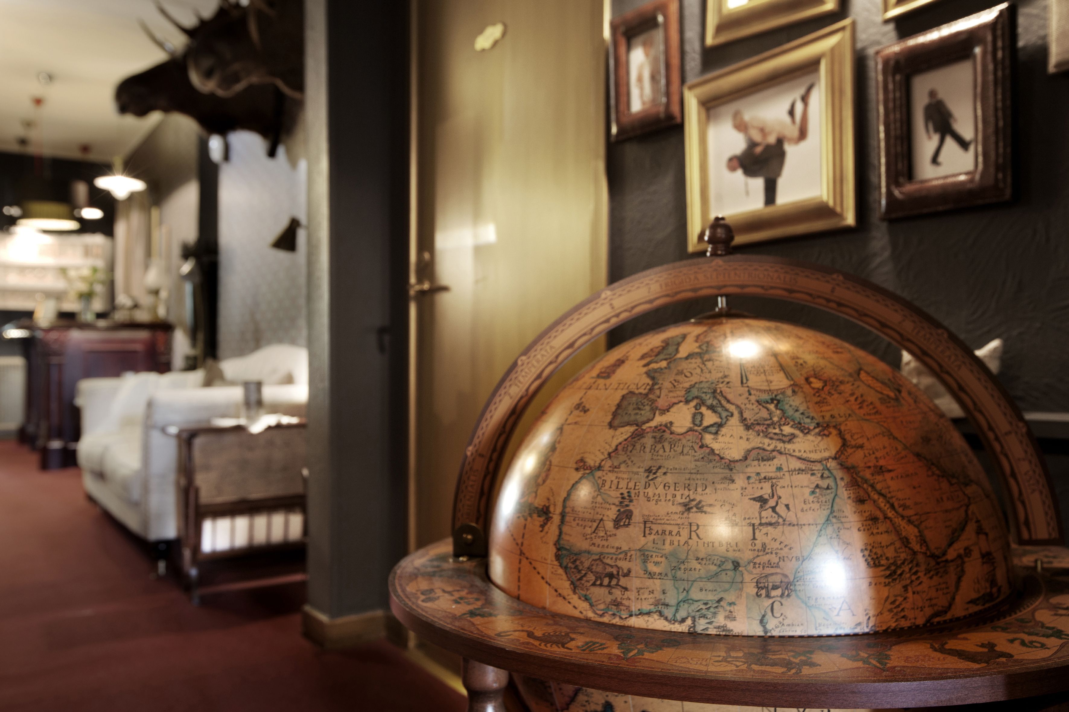 A globe on a wooden stand in a room with framed pictures on the wall and a sofa in the background.