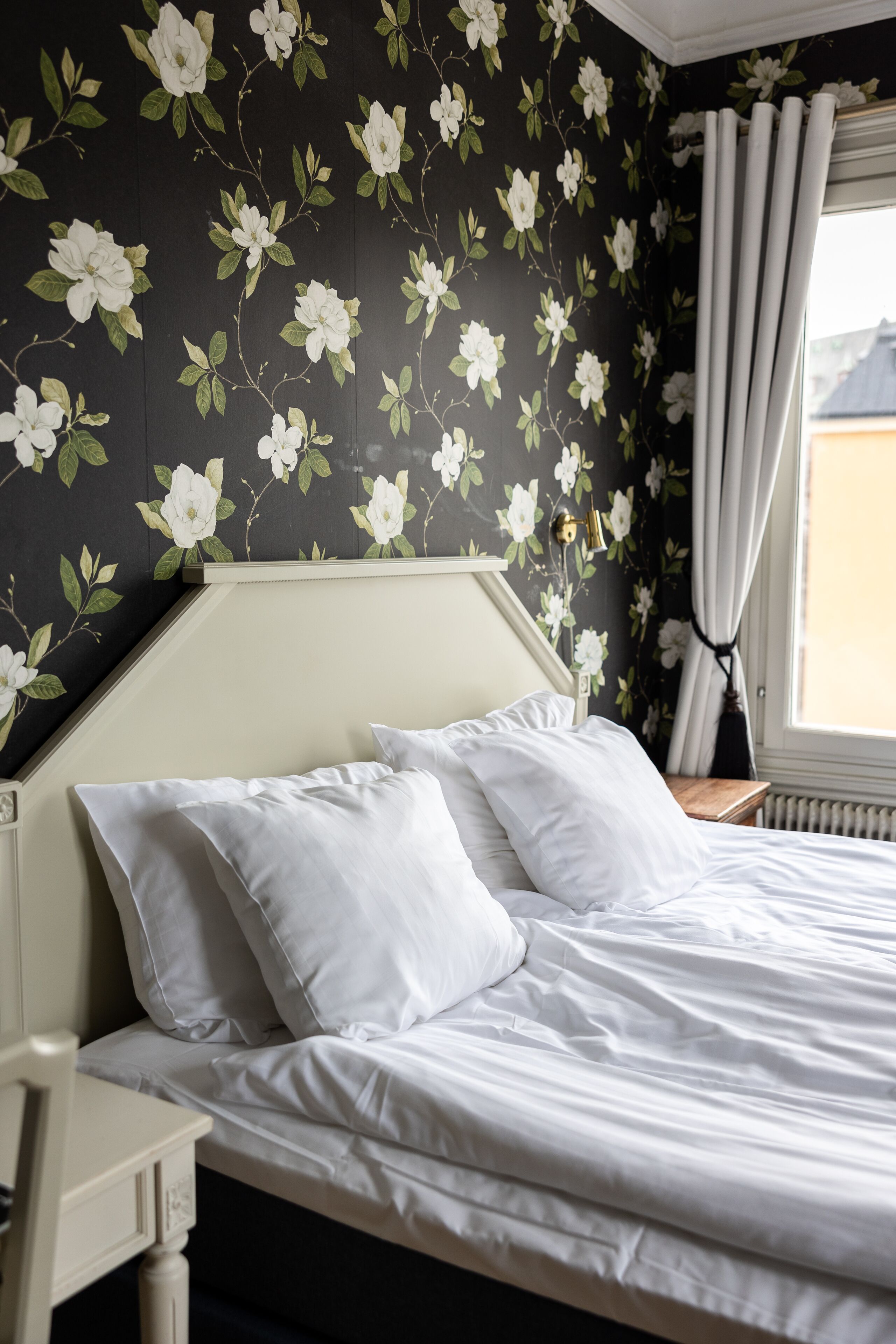 Una cama bien tendida con sábanas y almohadas blancas, una mesita de noche con una lámpara y un estampado floral en la pared.
